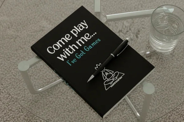 Black notebook with 'Come play with me... I've Got Games' text and a pen on a glass table.